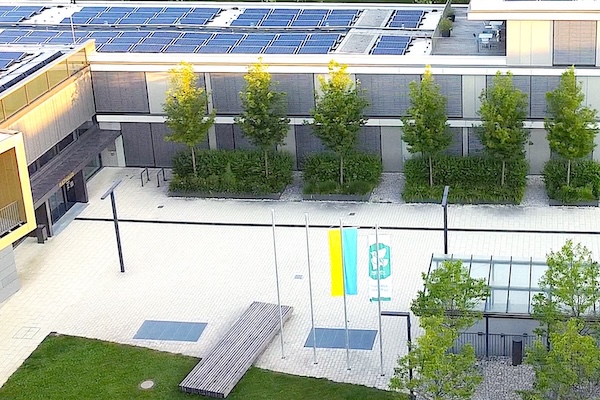 Luftbild mit seitlichem Blick auf den Rathaus-Vorplatz. Deutlich ist der helle Boden-Belag zu erkennen zwischen vielen grünen Bäumen und Rasen.